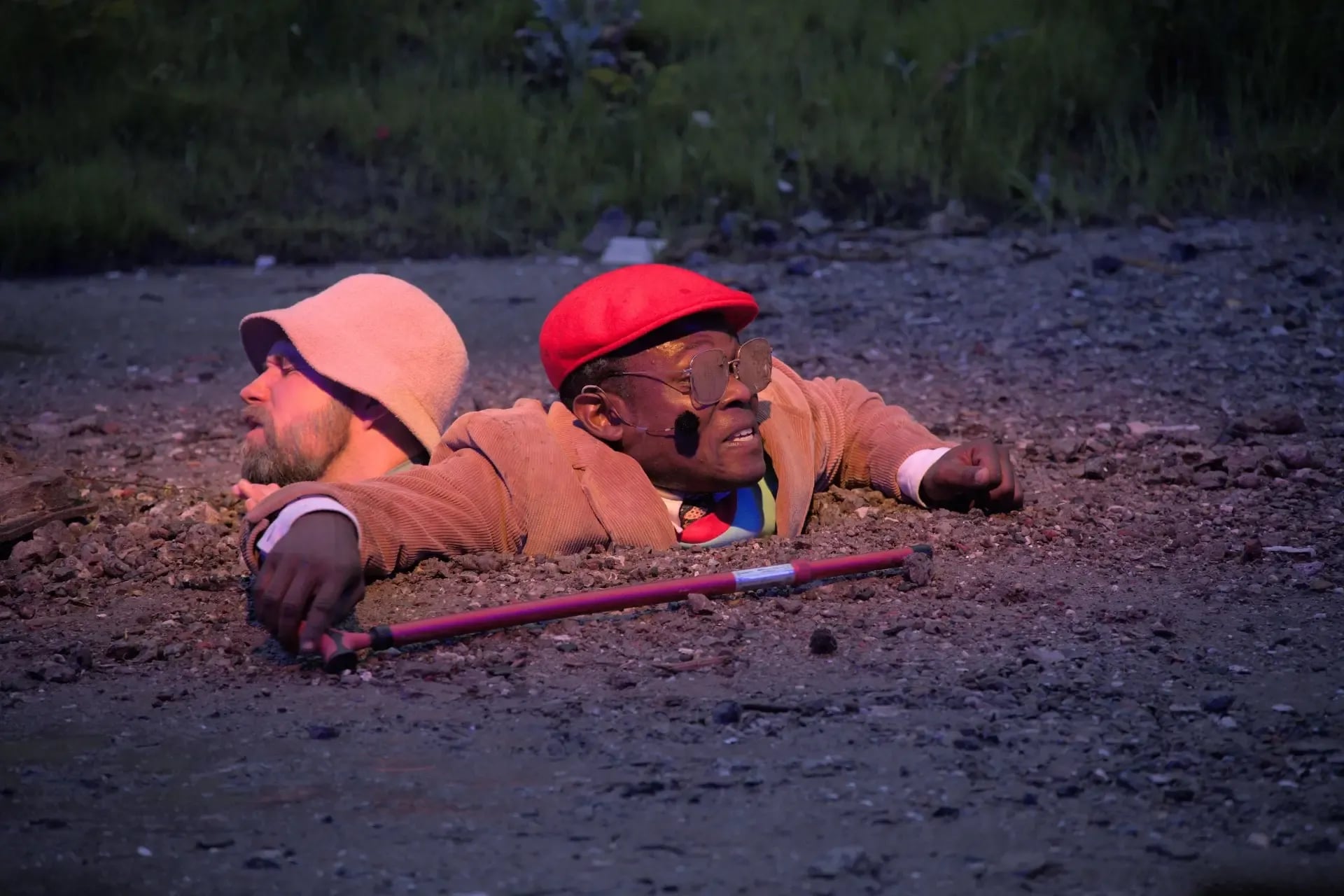 Twee mannen zitten tot hun nek vast in de grond op een grindachtig terrein; één draagt een rode pet en houdt een wandelstok vast, terwijl de ander een lichte hoed draagt en wegkijkt.